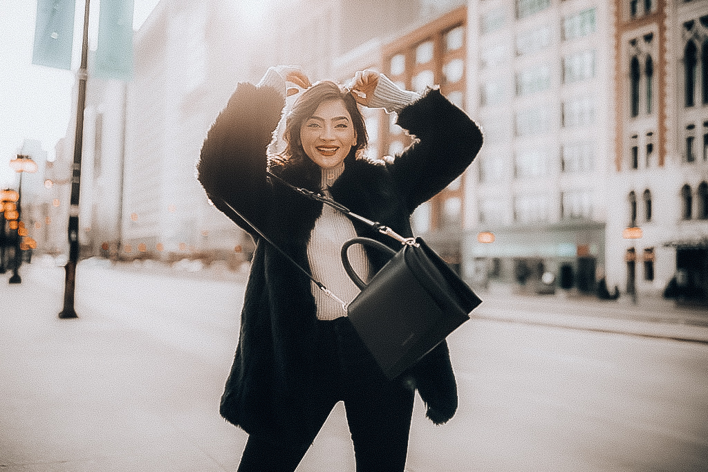 Bunette romanian blogger andreea cristina is smiling in downtown Chicago wearing a beige turtle neck, a gold necklace, a black faux fur jacket, highwaisted black jeans and mid calf black boots.