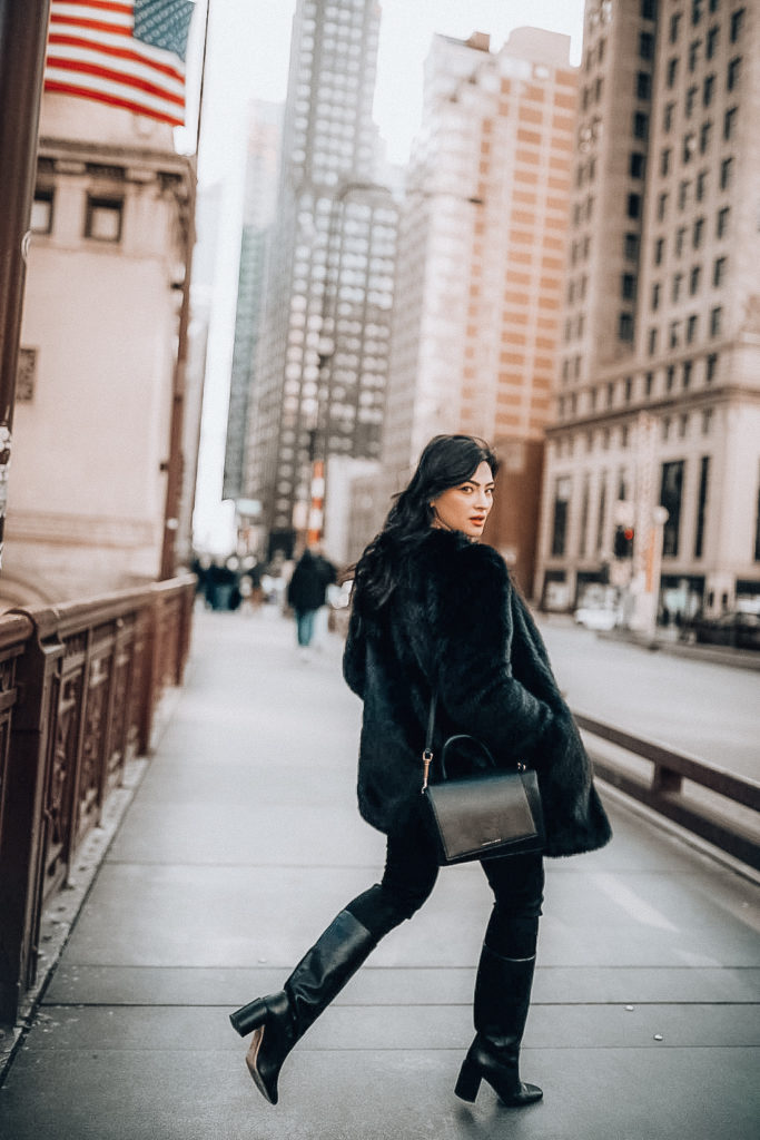 Bunette romanian blogger andreea cristina is walking in downtown Chicago wearing a beige turtle neck, a gold necklace, a black faux fur jacket, highwaisted black jeans and mid calf black boots.