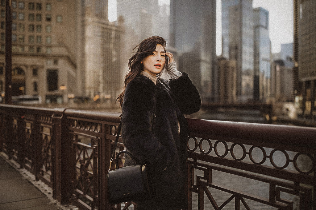 brunette fashion blogger (andreea cristina) standing on the chicago river walk in a black Talbots faux fur coat. She has a black back and long, wavy hair. The chicago city scape is in the background.